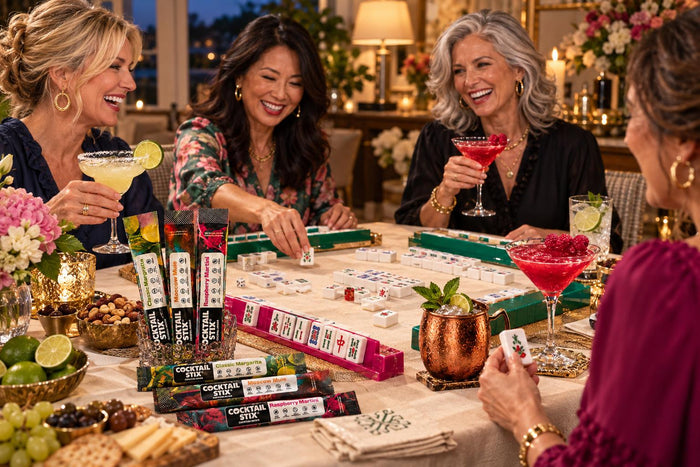 four adults playing American Mah Jongg with cocktails beside the table. Overhead view of tiles, racks, cards, coasters, and drinks.